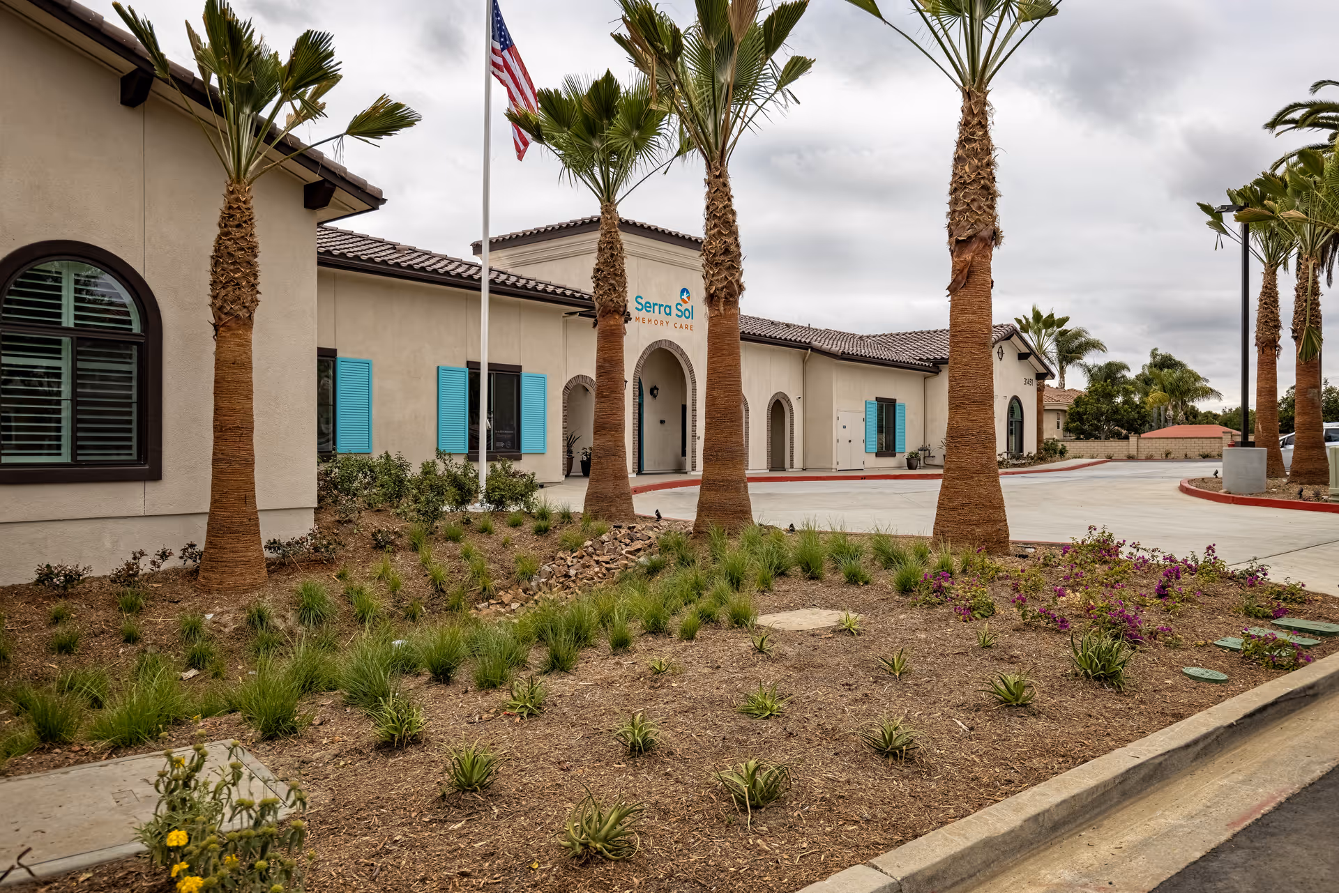 Exterior view of Serra Sol Memory Care facility showing a beige building with blue window shutters, several palm trees, landscaped garden beds with small plants and flowers, an American flag on a flagpole, and a cloudy sky.