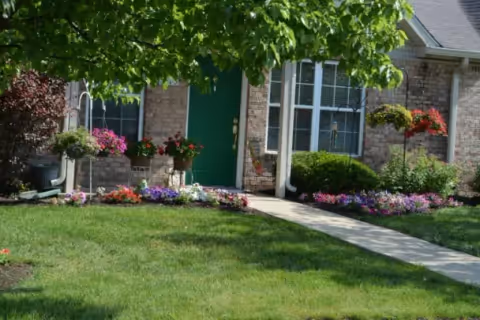 Front exterior view of a brick residential building with a green door, windows, a concrete walkway, and a well-maintained lawn and flower beds with colorful flowers and shrubs.