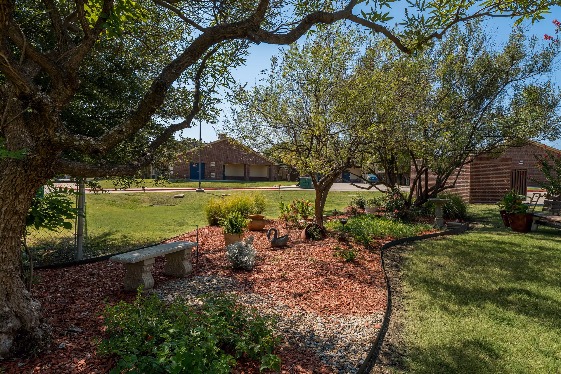 A landscaped garden area with a variety of plants, trees, and decorative elements including a stone bench, potted plants, and a swan sculpture. The garden is bordered by a curved edge separating mulch and grass. In the background, there are brick buildings under a clear blue sky.