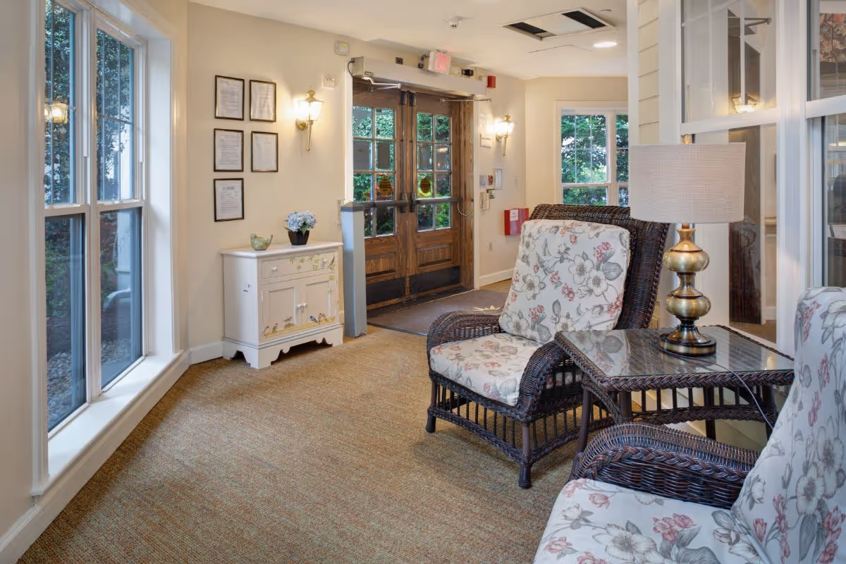 A cozy sitting area near the entrance of a facility with floral cushioned wicker chairs, a glass-top wicker side table with a brass lamp, a small white cabinet with decorative items, wall-mounted lights, and large windows letting in natural light.
