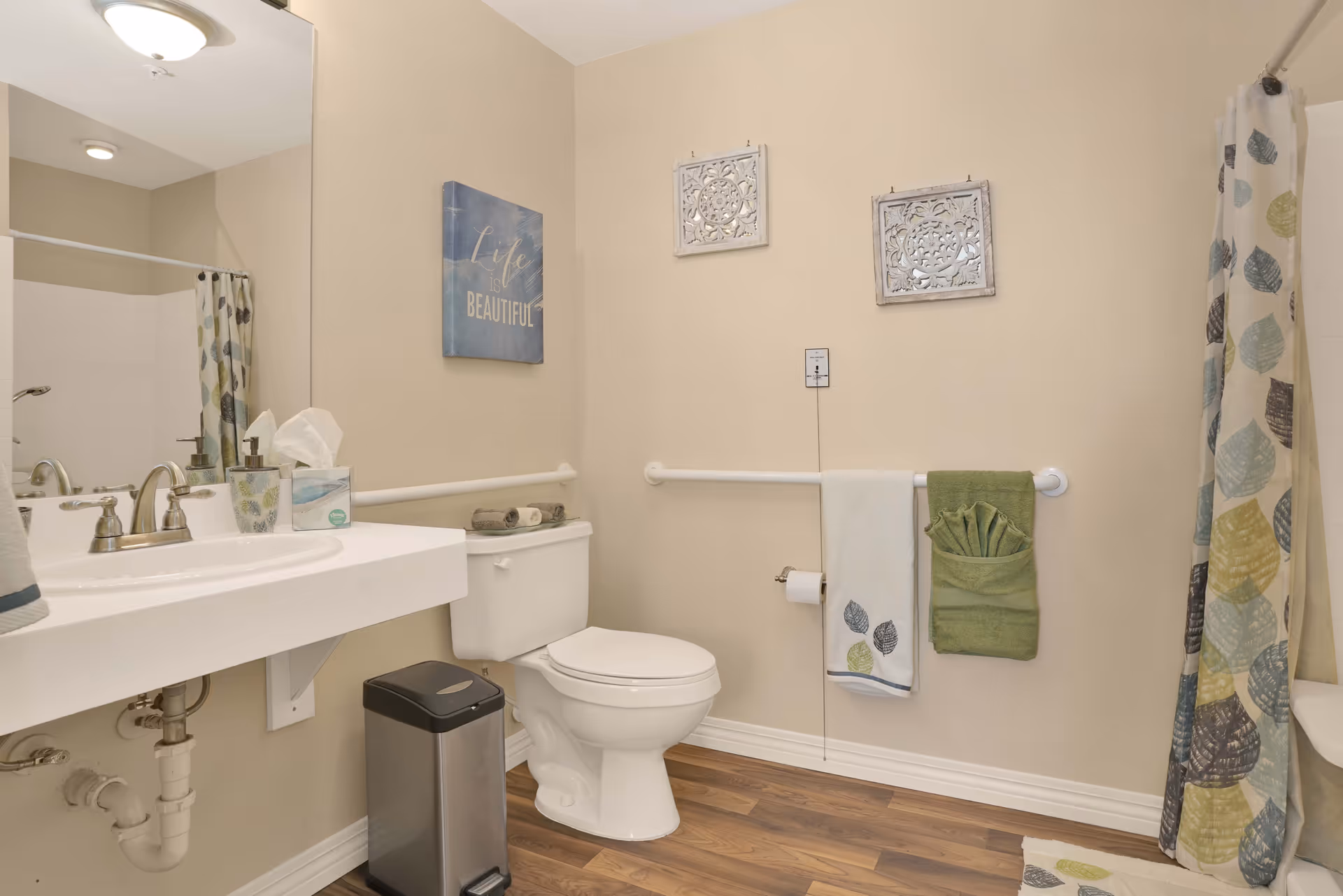A clean and accessible bathroom with a white toilet, a wall-mounted sink with a silver faucet, a trash can, and a shower with a curtain featuring a leaf pattern. The walls are beige with two decorative wall hangings and a sign that reads 'Life is Beautiful'. There are grab bars installed near the toilet and along the wall with towels hanging on them.