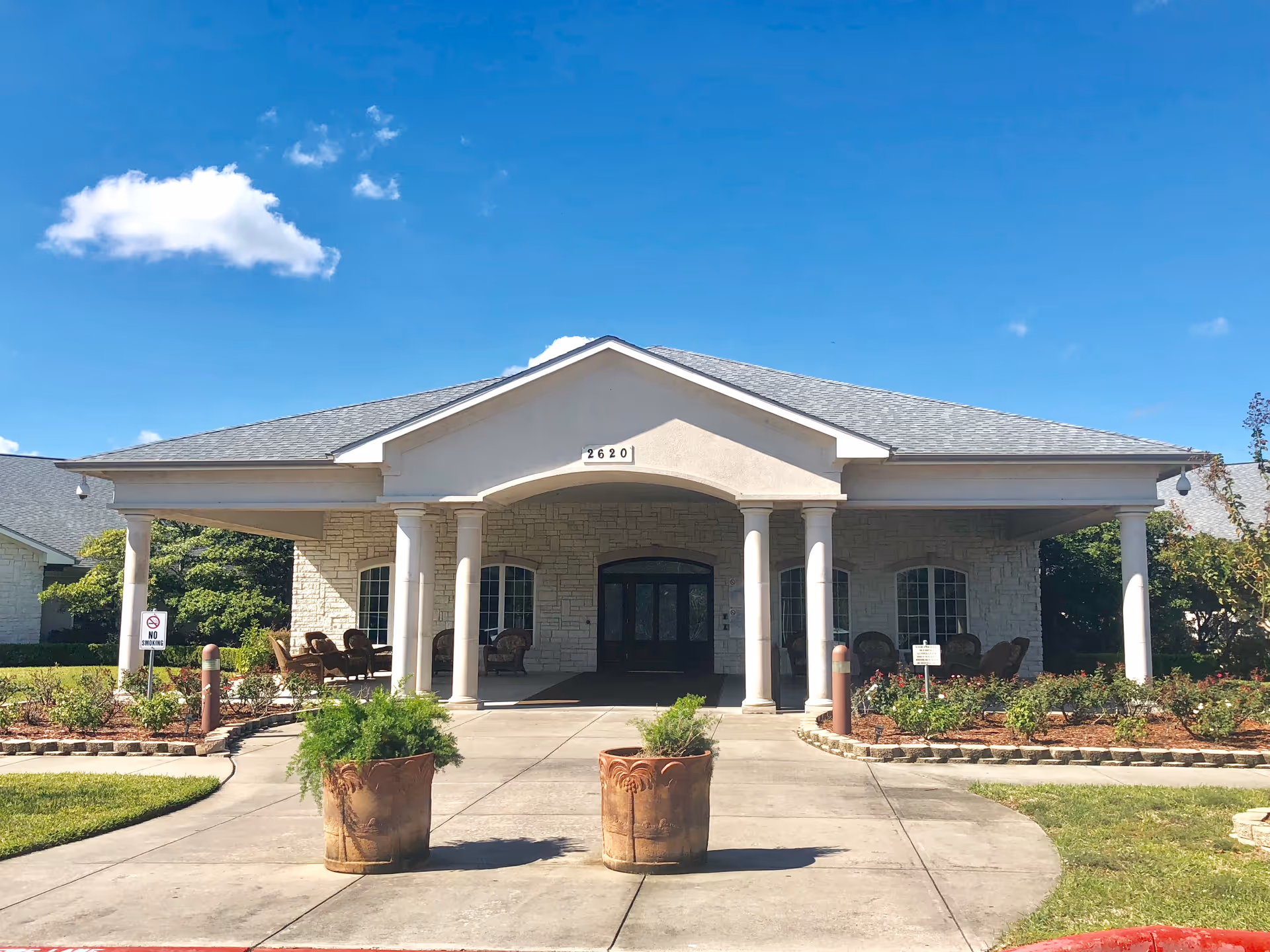 Front exterior view of a single-story building with a covered entrance supported by four white columns. The building has a stone facade and a gray shingled roof. Two large potted plants are placed on the concrete driveway leading to the entrance. There are chairs arranged on the porch area under the roof. The sky is clear with a few clouds.