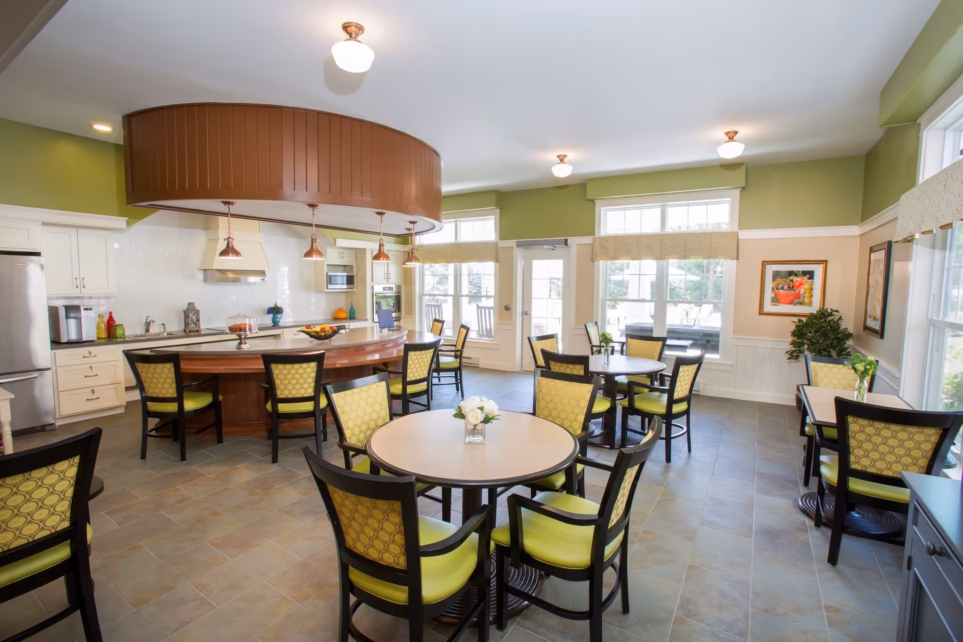 A bright dining area with multiple round tables and chairs featuring green cushions and patterned backs. The room has large windows allowing natural light, green walls with white trim, and a kitchen area with white cabinets, stainless steel appliances, and a curved wooden counter with pendant lights hanging above.