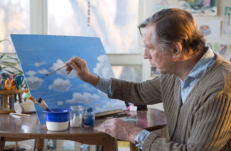 An elderly man painting white clouds on a blue sky canvas at a table indoors, with paint jars and brushes nearby.