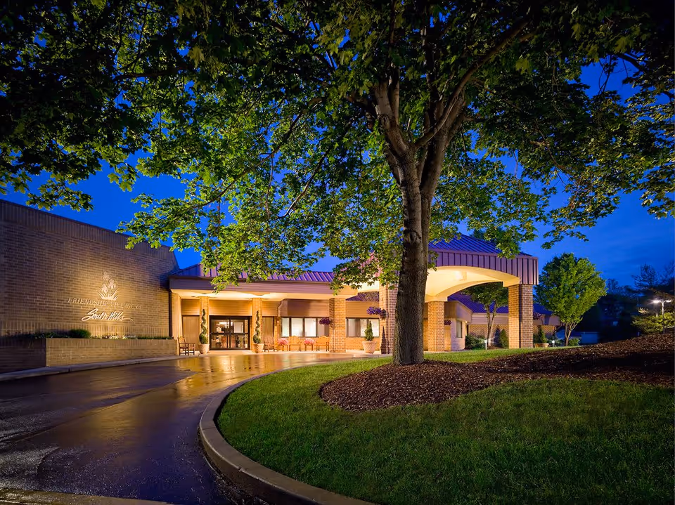 Evening exterior view of Friendship Village of South Hills building entrance with illuminated signage, a covered drop-off area, and surrounding trees and landscaping.