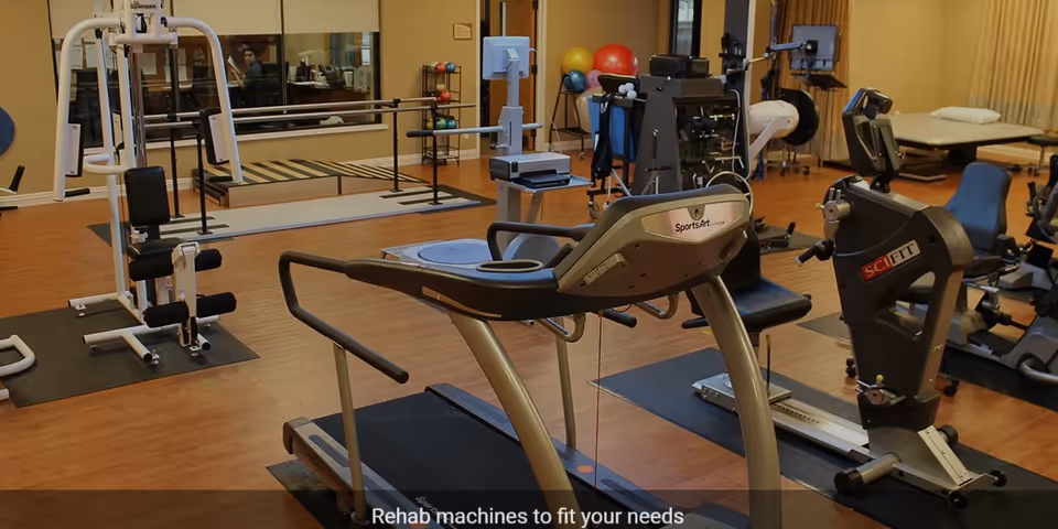 A rehabilitation gym room with various exercise and rehab machines including a treadmill, stationary bike, weight machine, and parallel bars. The room has wooden flooring and beige walls with some windows and a treatment table in the background.