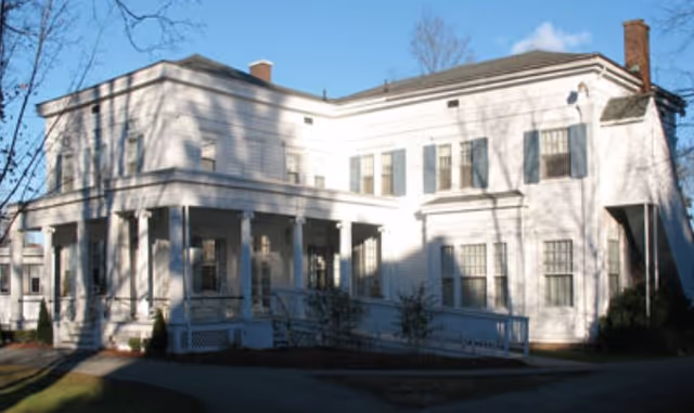 Exterior view of a large white two-story building with multiple windows and a covered porch with columns, surrounded by bare trees and a paved walkway.
