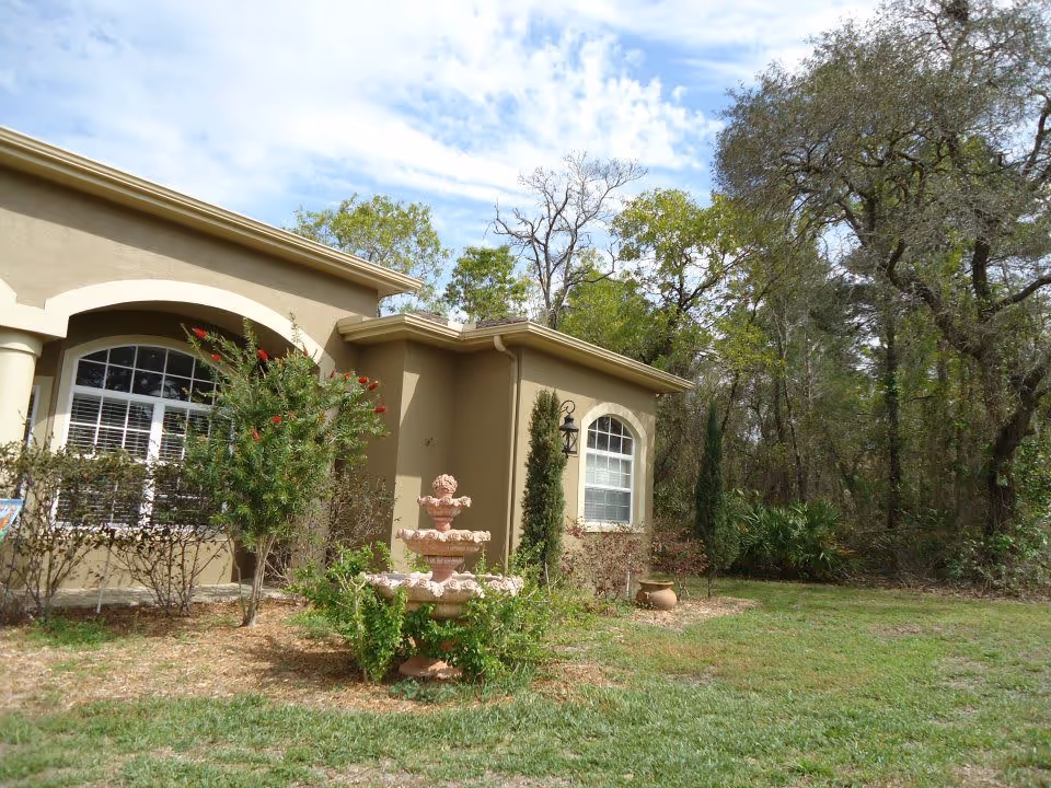 Exterior view of a beige single-story building with arched windows and a small garden in front featuring a tiered stone fountain surrounded by greenery. Trees and a partly cloudy sky are visible in the background.