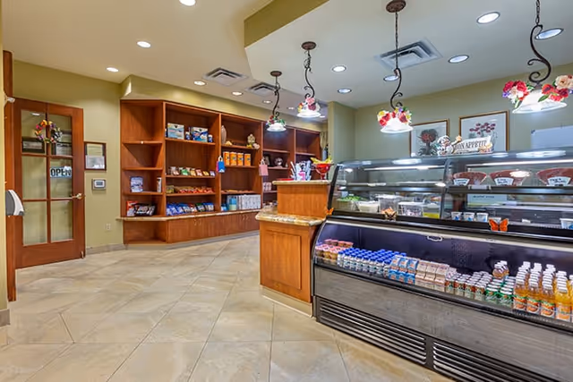Interior view of a small retail or snack area in a senior living facility with a refrigerated display case containing beverages and snacks, wooden shelves stocked with various packaged goods, and decorative hanging lights with floral designs.