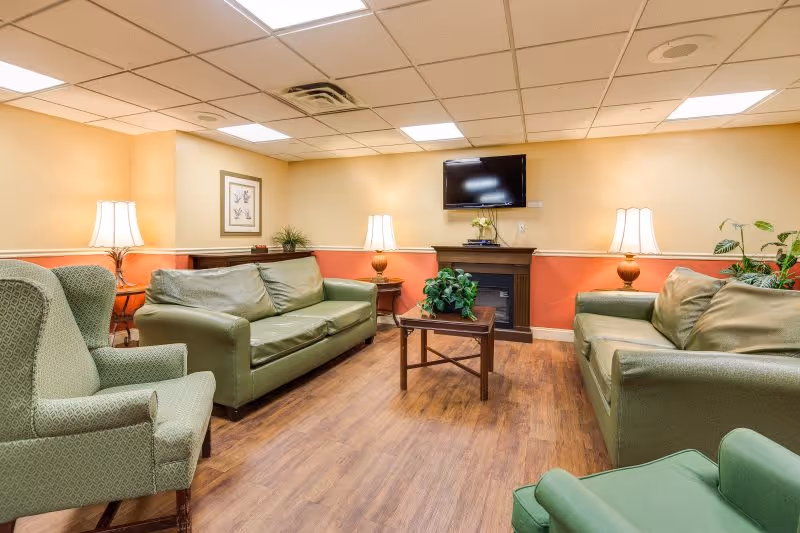 A cozy senior living facility lounge with green upholstered sofas and armchairs arranged around a small wooden coffee table with a plant. The room has warm beige and coral walls, wooden flooring, two table lamps on side tables, a framed picture on the wall, a flat-screen TV mounted above an electric fireplace, and ceiling tiles with recessed lighting.