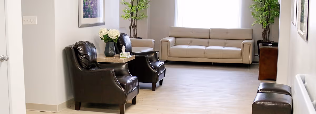 A bright and clean living room area with two black leather armchairs and a small table with a vase of flowers and a tissue box between them. In the background, there is a beige sofa, a wooden side table with a plant, and a large window letting in natural light. The floor is light-colored wood, and the walls are painted white with framed artwork.