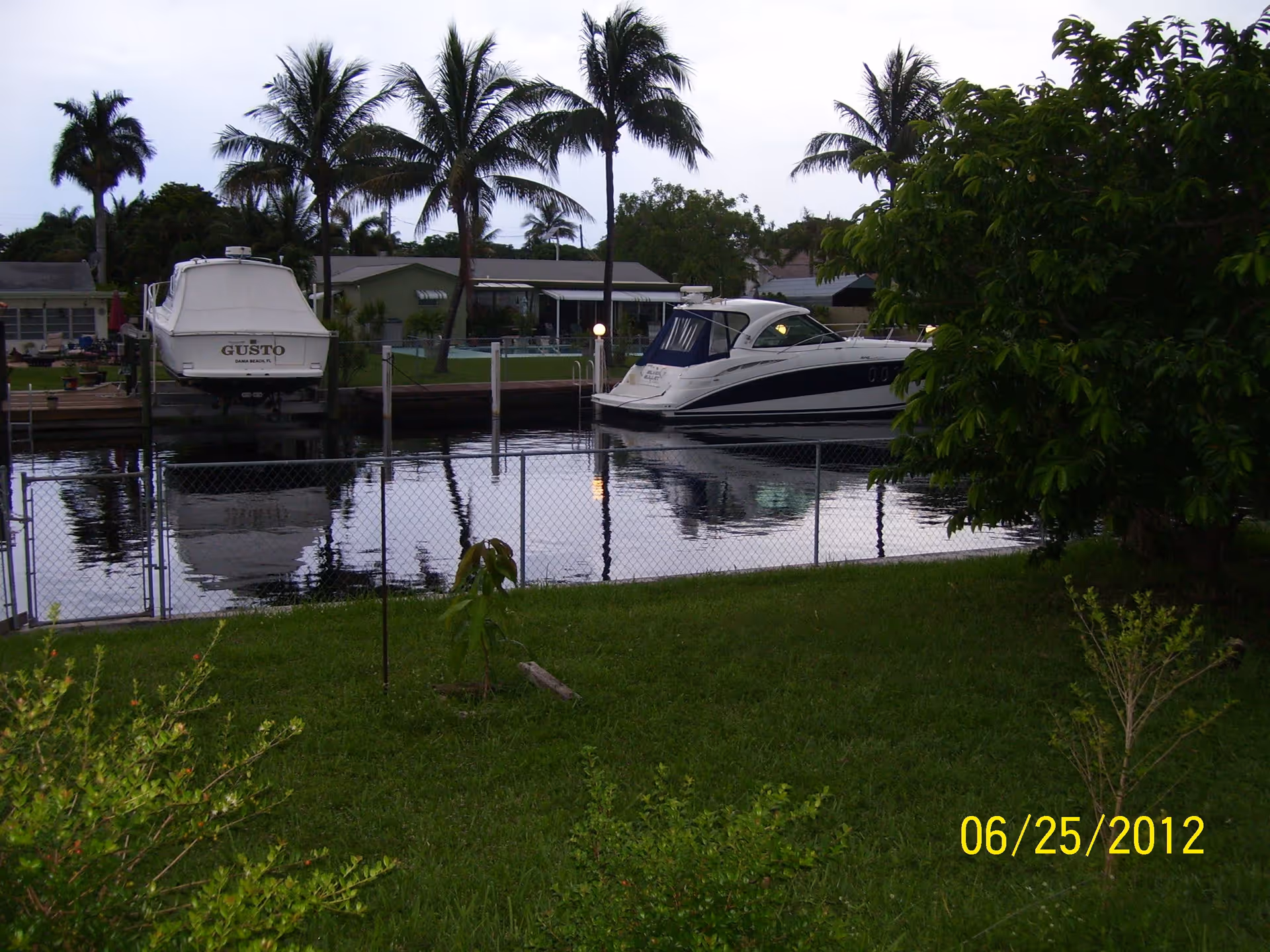 A waterfront view showing two boats docked near a residential area with palm trees and green grass in the foreground. There is a chain-link fence separating the grassy area from the water. The sky is overcast.