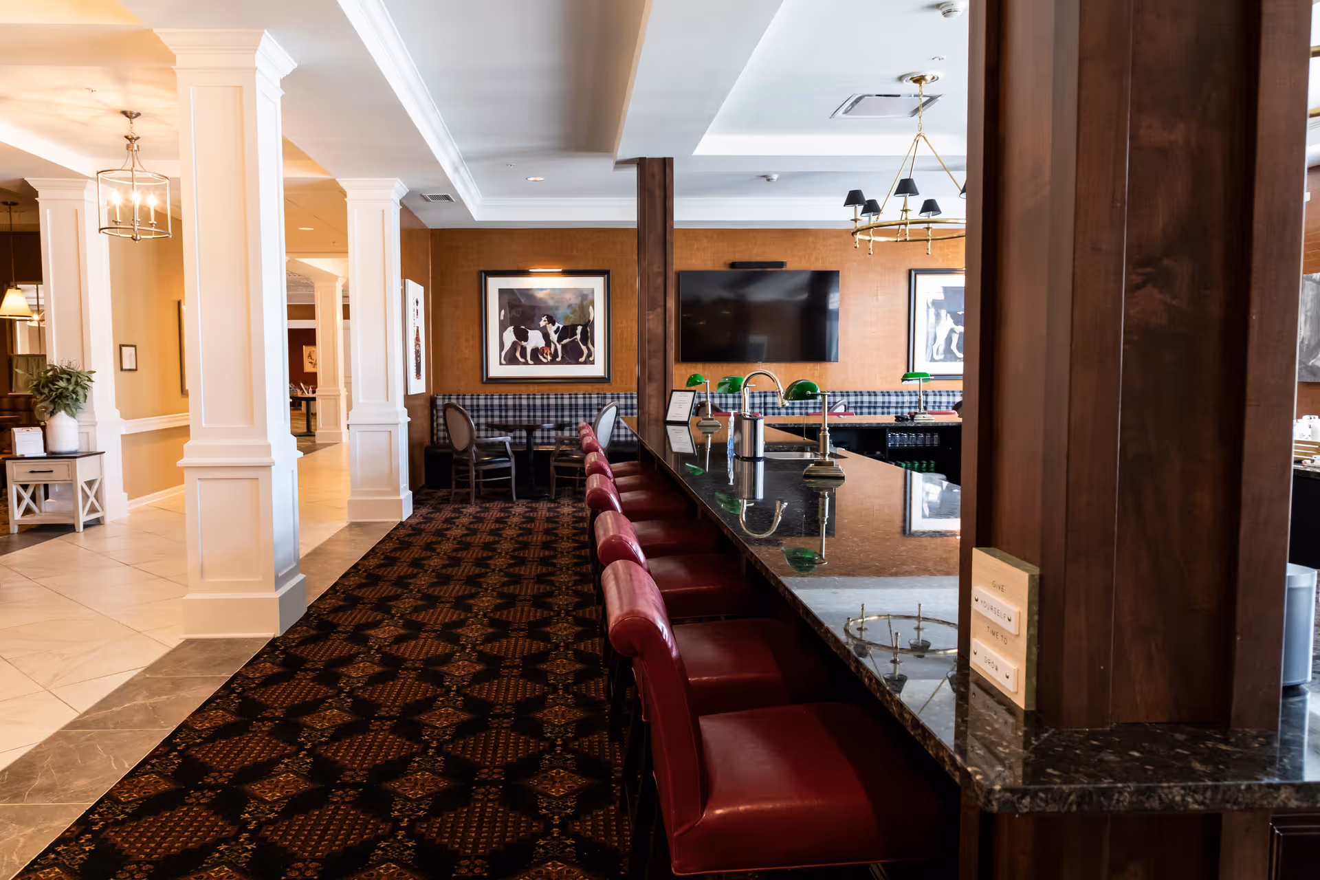 Interior view of a senior living facility lounge area with a long granite countertop bar and red cushioned bar stools. The room features patterned carpet, white columns, framed artwork on the walls, a flat-screen TV, and a seating area with plaid upholstered chairs and tables. Warm lighting fixtures hang from the ceiling.