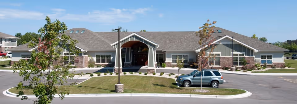 Front exterior view of a single-story building with a gabled roof, brick and light-colored siding, a covered entrance, and a small parking area with a silver SUV parked. The building is surrounded by a well-maintained lawn and a few young trees under a partly cloudy blue sky.