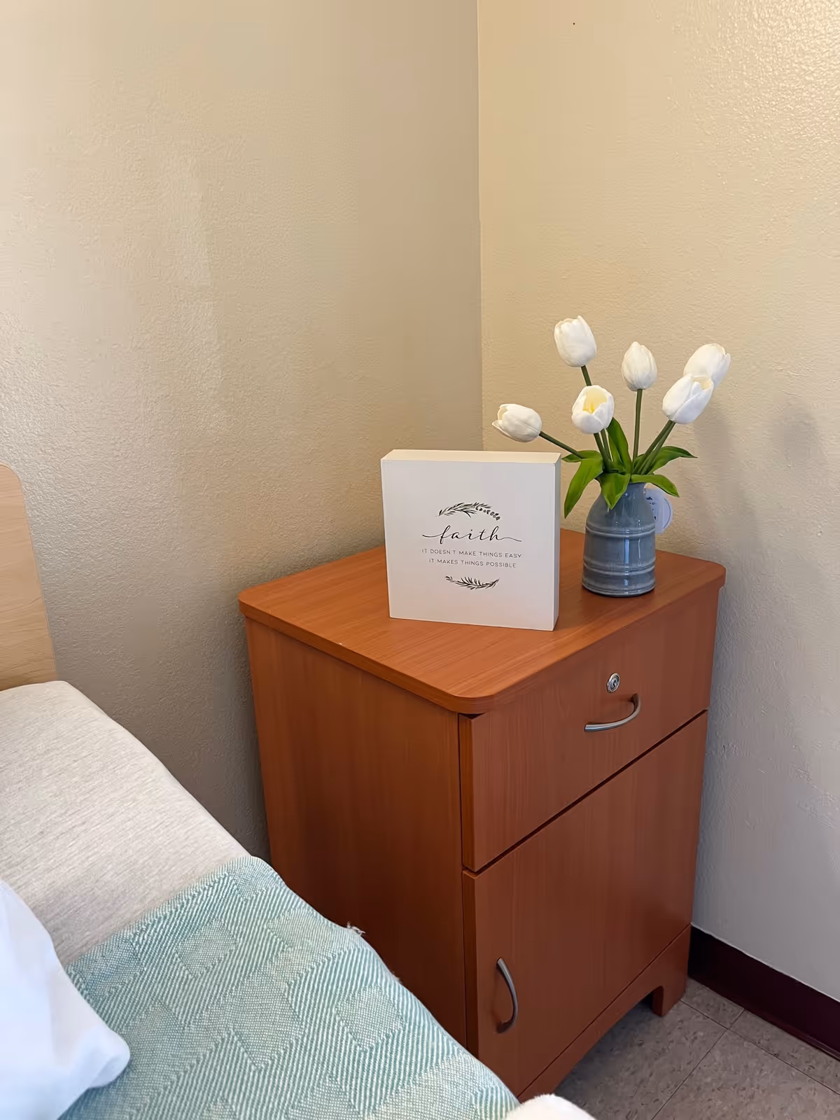 A wooden bedside table next to a bed with a light green patterned blanket. On the table, there is a small decorative sign with the word 'faith' and a quote, along with a gray vase holding white tulips. The walls are beige and the floor has light-colored tiles.