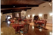 A spacious dining room with wooden floors and ceiling beams, featuring multiple round tables covered with red tablecloths and set with glassware and napkins. The room is softly lit with chandeliers and wall sconces, and there is a stone fireplace at the far end.