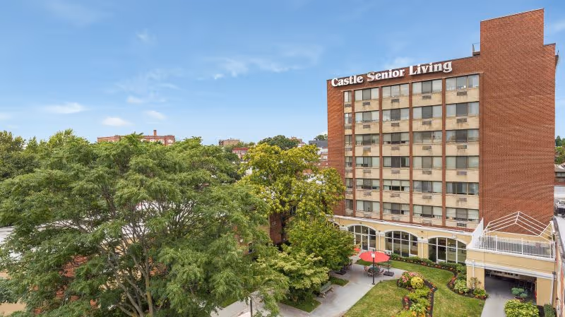 Exterior view of a multi-story brick building with the sign 'Castle Senior Living' on top. The building is surrounded by trees and landscaped greenery, with a small patio area featuring a red umbrella and seating. The sky is clear with a few clouds.