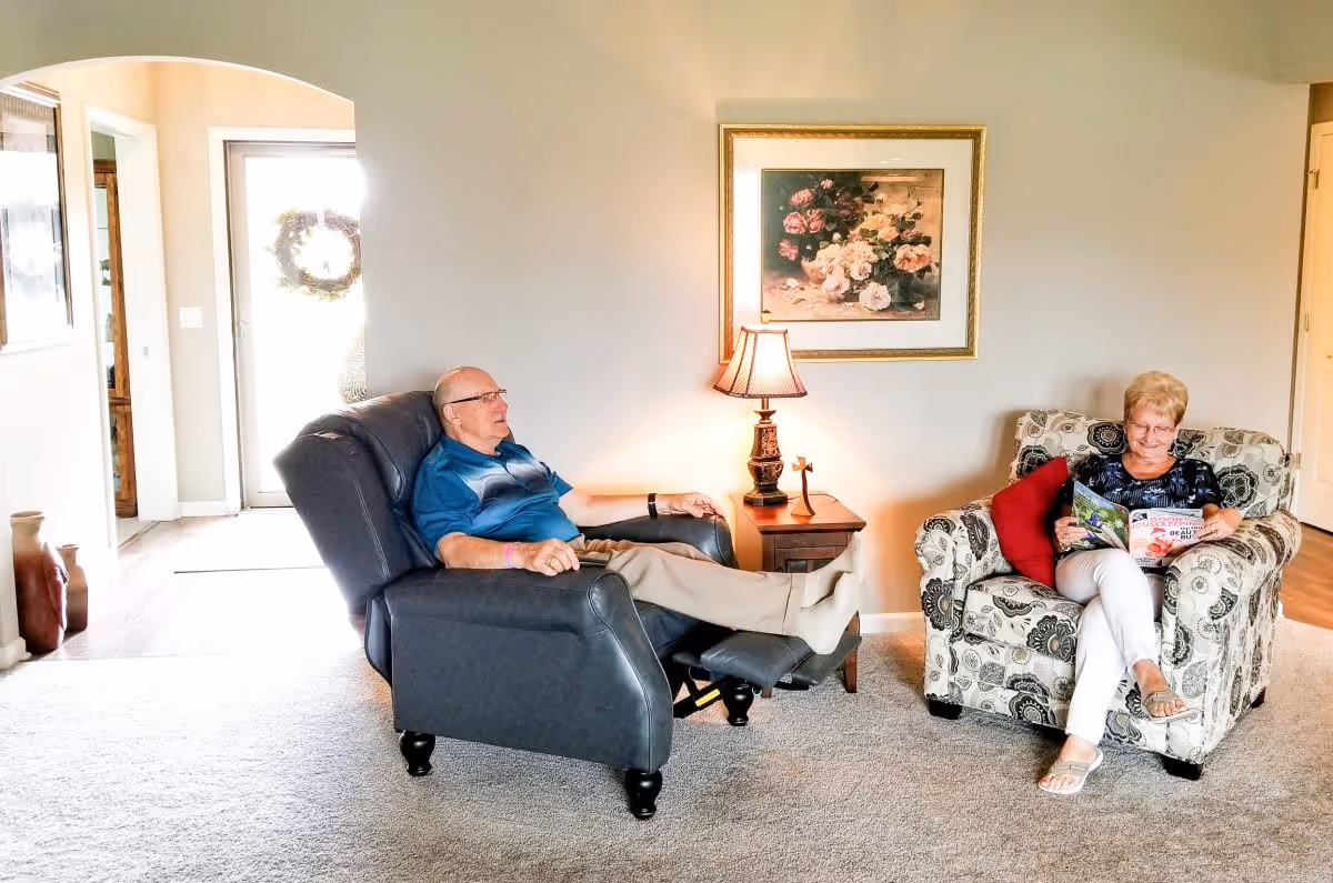 An elderly man relaxing in a black recliner chair with his feet up, and an elderly woman sitting in a patterned armchair reading a magazine in a cozy living room. There is a side table with a lamp and a small cross between them, a framed floral painting on the wall, and a bright doorway in the background.