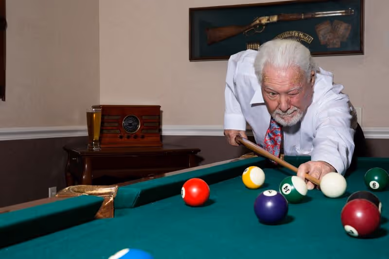 An elderly man with white hair and a beard is playing pool, aiming to strike the cue ball on a green pool table. Behind him, there is a wooden table with a vintage radio and a glass of beer. A framed display of a rifle and some bullets hangs on the wall.