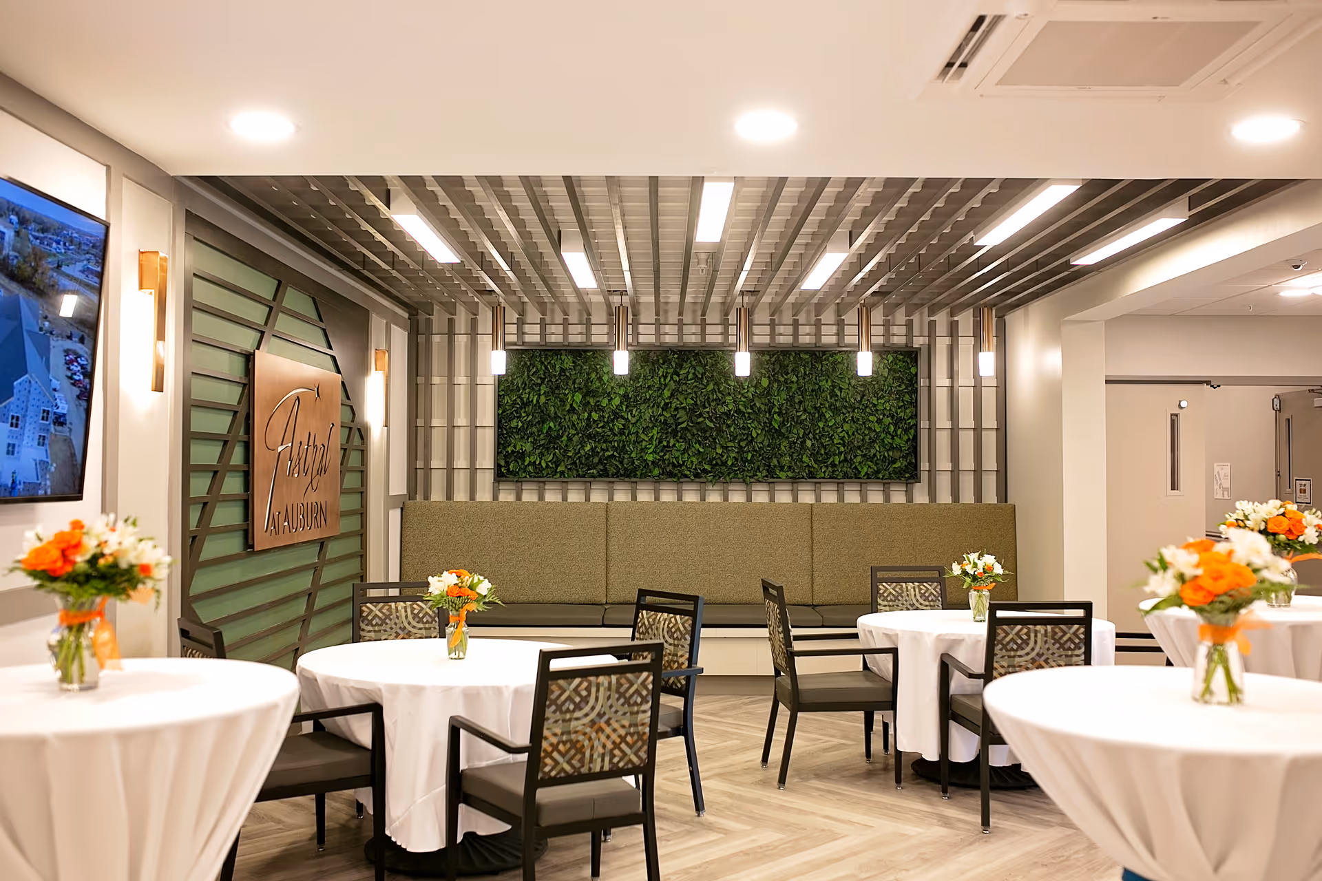 Interior view of a dining area at Astral at Auburn featuring round tables covered with white tablecloths, each adorned with a small vase of orange and white flowers. The room has modern lighting fixtures hanging from a decorative ceiling, a green plant wall, and a cushioned bench along the back wall. A sign with the facility name is mounted on the left wall.