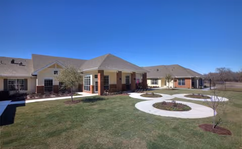 Exterior view of a single-story senior living facility building with beige walls, brick accents, and a gray shingled roof. The building surrounds a landscaped courtyard with green grass, small trees, and circular garden beds under a clear blue sky.