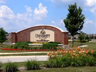A large brick and stone sign for University Place, a Continuing Care Retirement Community (CCRC), surrounded by well-maintained landscaping including flowers, shrubs, and a tree, with a partly cloudy blue sky in the background.