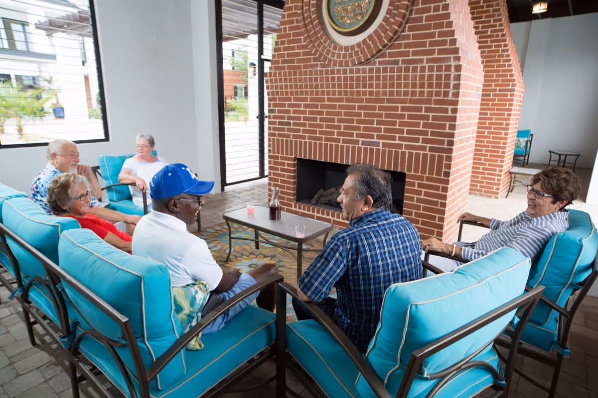 A group of six elderly people sitting on turquoise cushioned chairs arranged around a small table with drinks, in front of a large brick fireplace inside a bright room with large windows.
