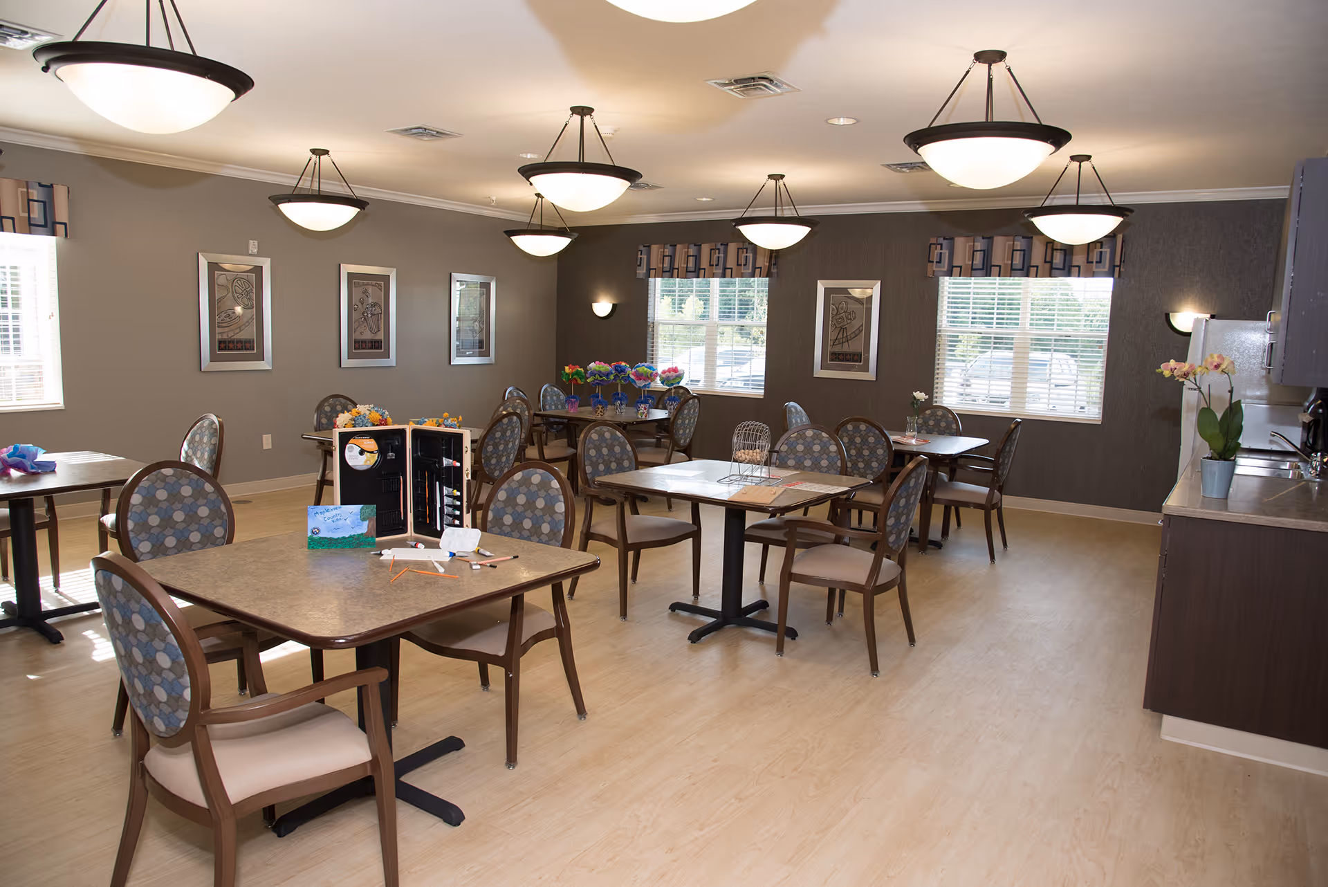 A well-lit dining room with multiple tables and chairs arranged neatly. The room has large windows with blinds and patterned valances, framed artwork on the walls, and several ceiling light fixtures. There is a kitchenette area with cabinets, a refrigerator, and a countertop on the right side. Some tables have decorative items like flowers and menus.