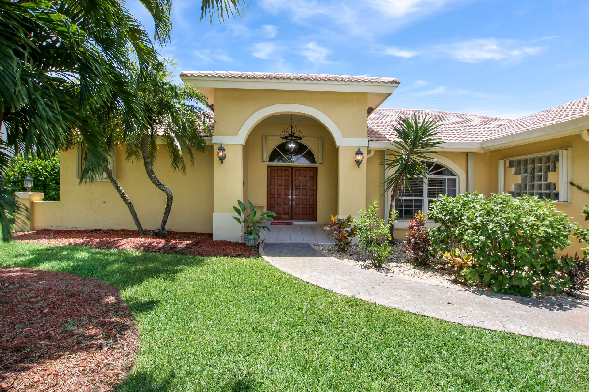 Front exterior view of a single-story building with a tiled roof and an arched entrance. The entrance has double wooden doors, two wall-mounted lanterns, and a hanging light fixture. The building is surrounded by green grass, palm trees, and various shrubs under a clear blue sky.