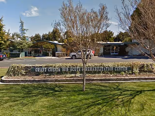 Front exterior view of Grant Cuesta Sub-acute and Rehabilitation Center with a sign in front, trees, parked cars, and a clear sky.