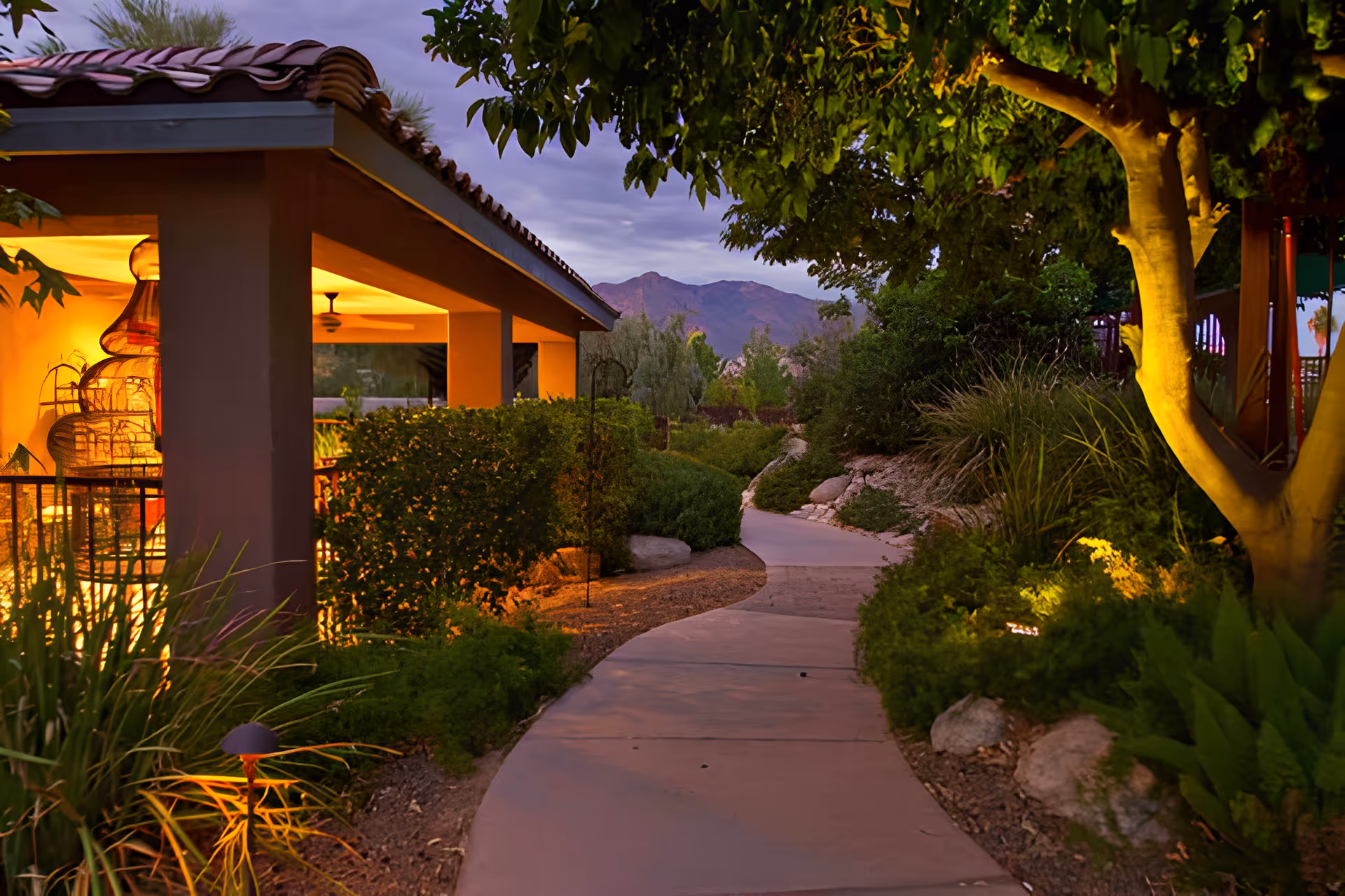 A winding outdoor pathway surrounded by lush greenery and trees, leading towards distant mountains under a twilight sky. On the left side, there is a covered patio area with warm yellow lighting and ceiling fans.
