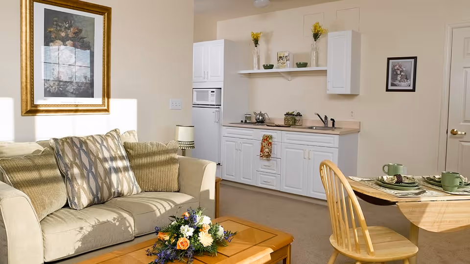 A cozy living area with a beige sofa adorned with patterned and solid cushions, a wooden coffee table with a floral arrangement, a small dining table set with green cups and plates, and a compact kitchen with white cabinets, a microwave, and decorative vases on a shelf.