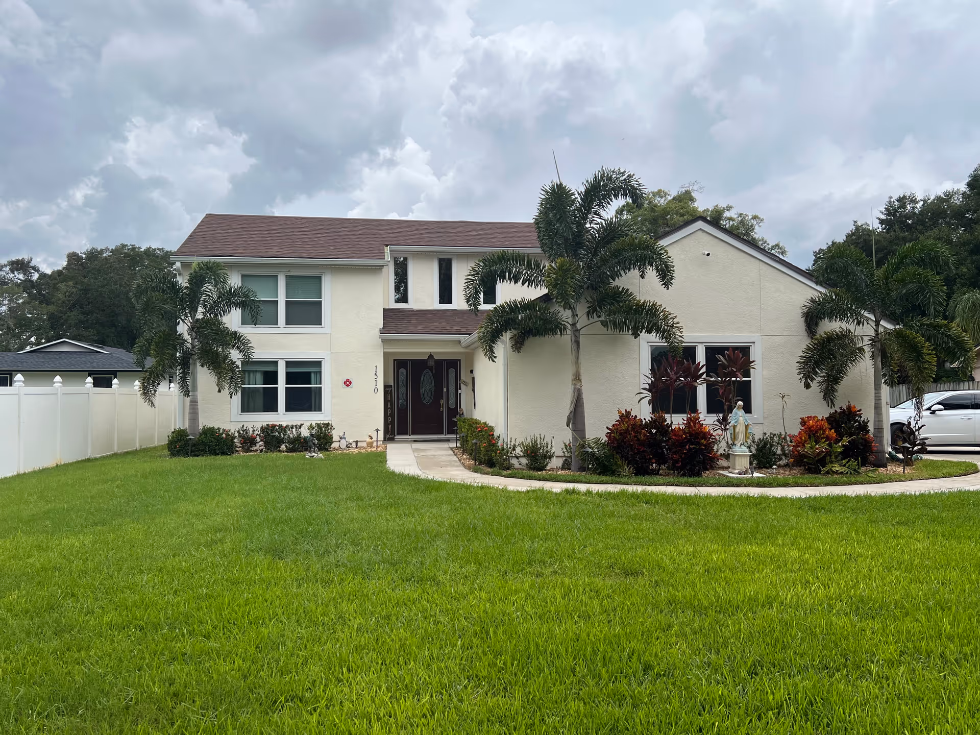 Front exterior view of a two-story assisted living facility building with a well-maintained green lawn, palm trees, and shrubs. The building has a light-colored facade with multiple windows and a central entrance with double doors. The sky is cloudy.