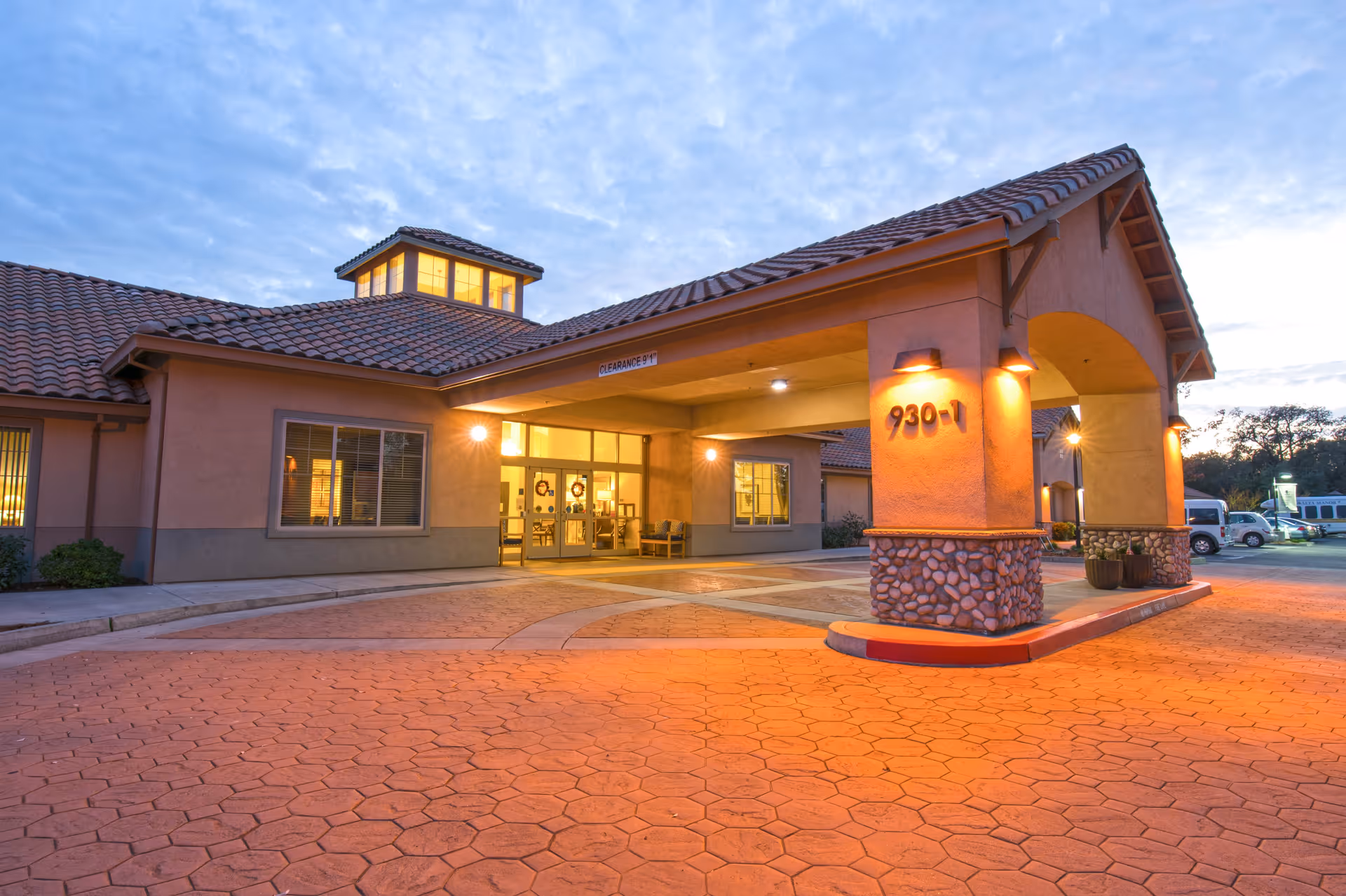 Exterior view of the entrance to Meadow Oaks of Roseville facility during dusk, showing a covered drop-off area with warm lighting, tiled roof, and a paved driveway.