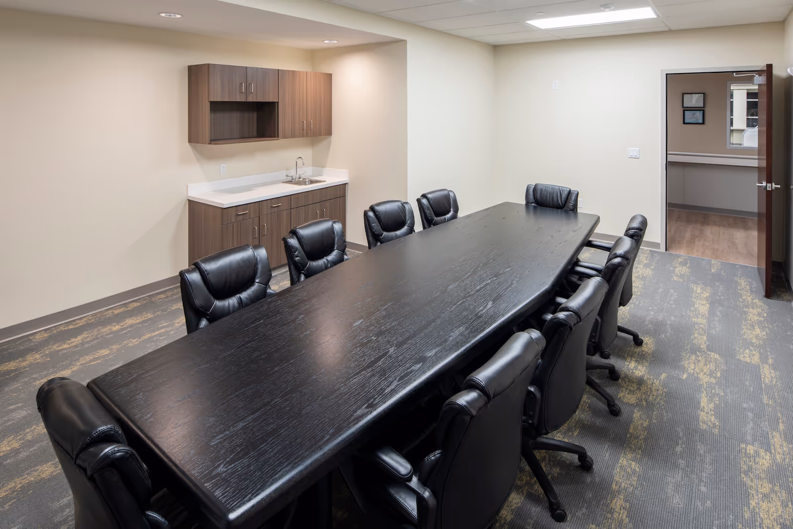 A conference room with a long black rectangular table surrounded by ten black leather office chairs. The room has beige walls, a carpeted floor with a patterned design, and a small kitchenette area with a sink and wooden cabinets. An open door leads to another room with wood flooring and framed pictures on the wall.