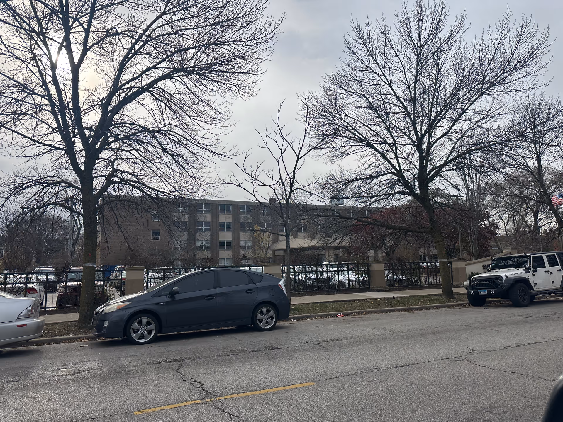 Street view of a multi-story brick building behind a black metal fence with several leafless trees in front. Several parked cars line the street, and the sky is overcast.