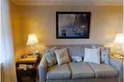 A cozy living room area with a gray sofa adorned with several patterned and plain cushions. On either side of the sofa are wooden side tables, each with a lamp emitting warm light. Above the sofa hangs a framed painting depicting boats on water with trees in the background.