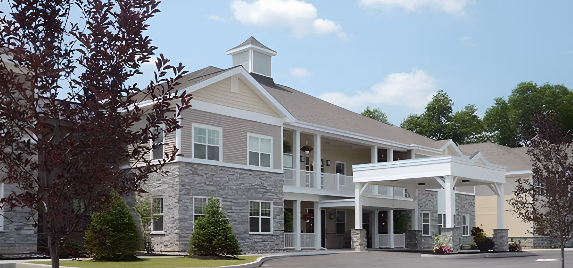 Exterior view of a senior living facility building with stone and beige siding, white trim, and a covered entrance. The building has two stories with multiple windows and a small tower-like structure on the roof. There are trees and shrubs around the building under a partly cloudy sky.