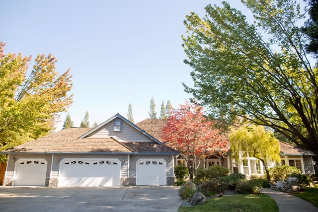 Front exterior view of a single-story senior living facility building with a three-car garage, surrounded by trees with green and red leaves under a clear sky.