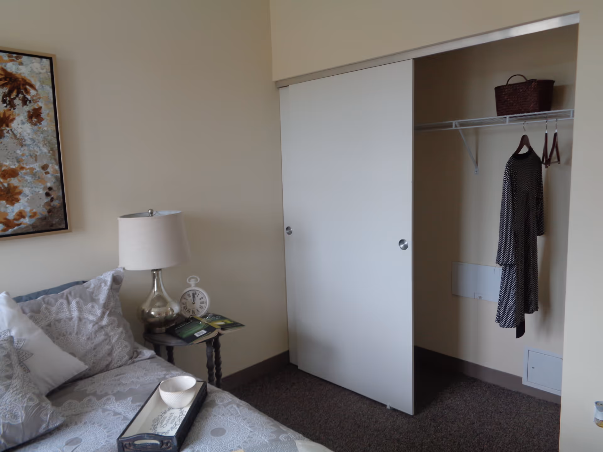 A bedroom corner with a bed featuring patterned bedding and pillows, a small round side table with a silver lamp, a white clock, and some magazines. Next to the bed is a closet with sliding white doors, one door open revealing a hanging black and white dress and a brown basket on the shelf above.