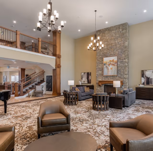 A spacious senior living facility common area with a large stone fireplace, two chandeliers, multiple seating arrangements including sofas and armchairs, a patterned carpet, and a staircase leading to an upper level with wooden railings.