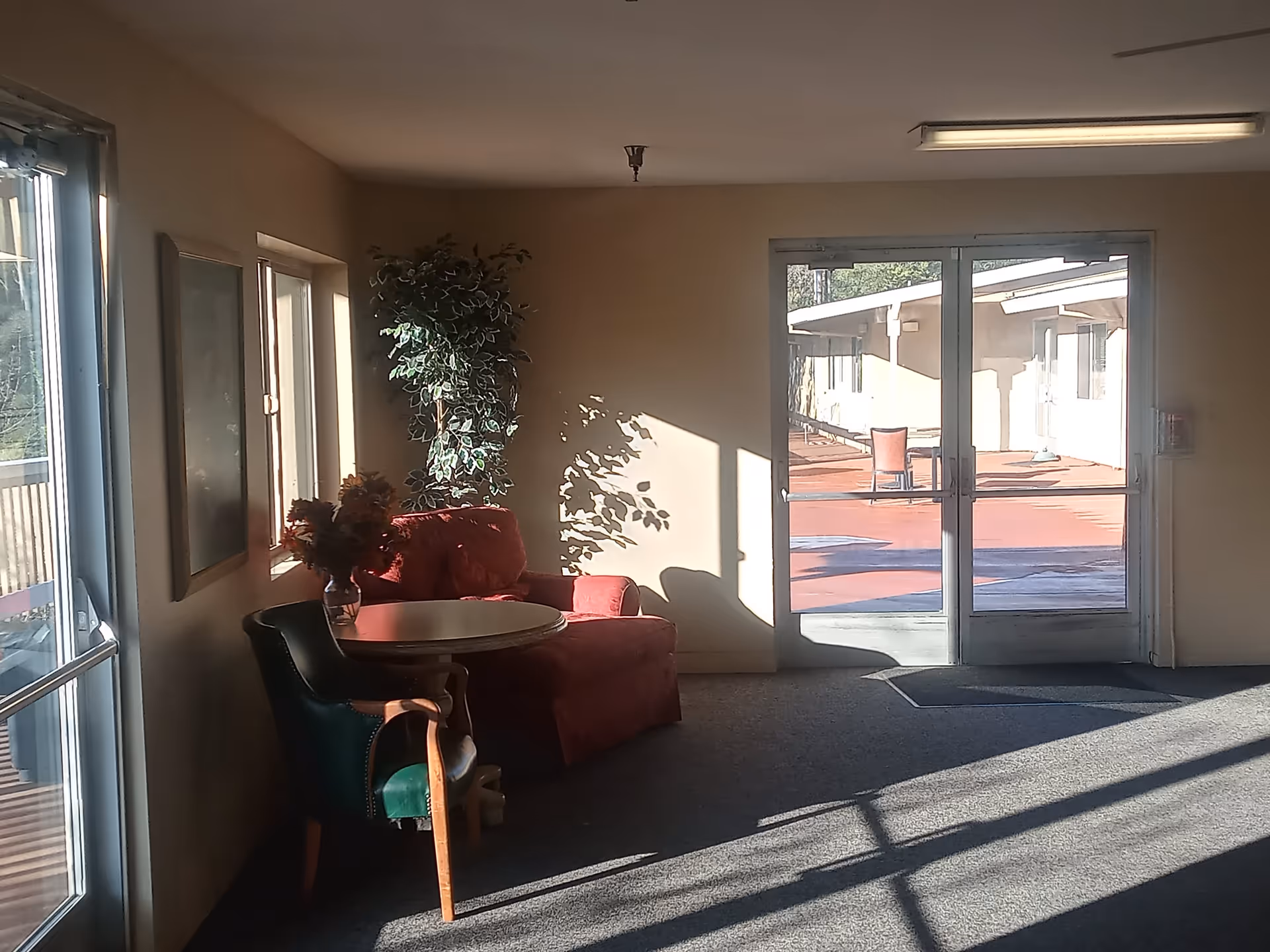 A sunlit corner of a senior living facility with a round wooden table, a green upholstered chair, a red armchair, a vase with flowers on the table, and a tall potted plant in the corner. Glass double doors lead to an outdoor patio area with another chair and table visible outside.