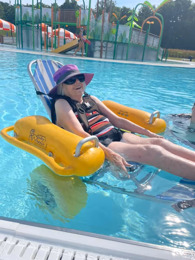 An elderly woman wearing a purple sunhat and sunglasses is relaxing in a pool on a yellow floating chair with armrests. She is smiling and wearing a striped swimsuit. In the background, there is a water play structure with slides and a child walking nearby.