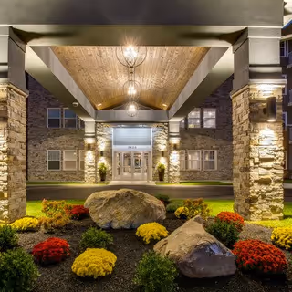 Entrance of a residential building at night with a covered driveway supported by stone pillars, illuminated by hanging lights. The landscaped area in front features large rocks and colorful flowers including red, yellow, and green plants.