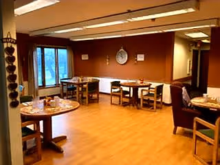 A dining area with several wooden tables and chairs arranged on a light wood floor. The walls are painted brown, and there is a large window letting in natural light. The room has a cozy and warm atmosphere with simple decor including a clock on the wall.