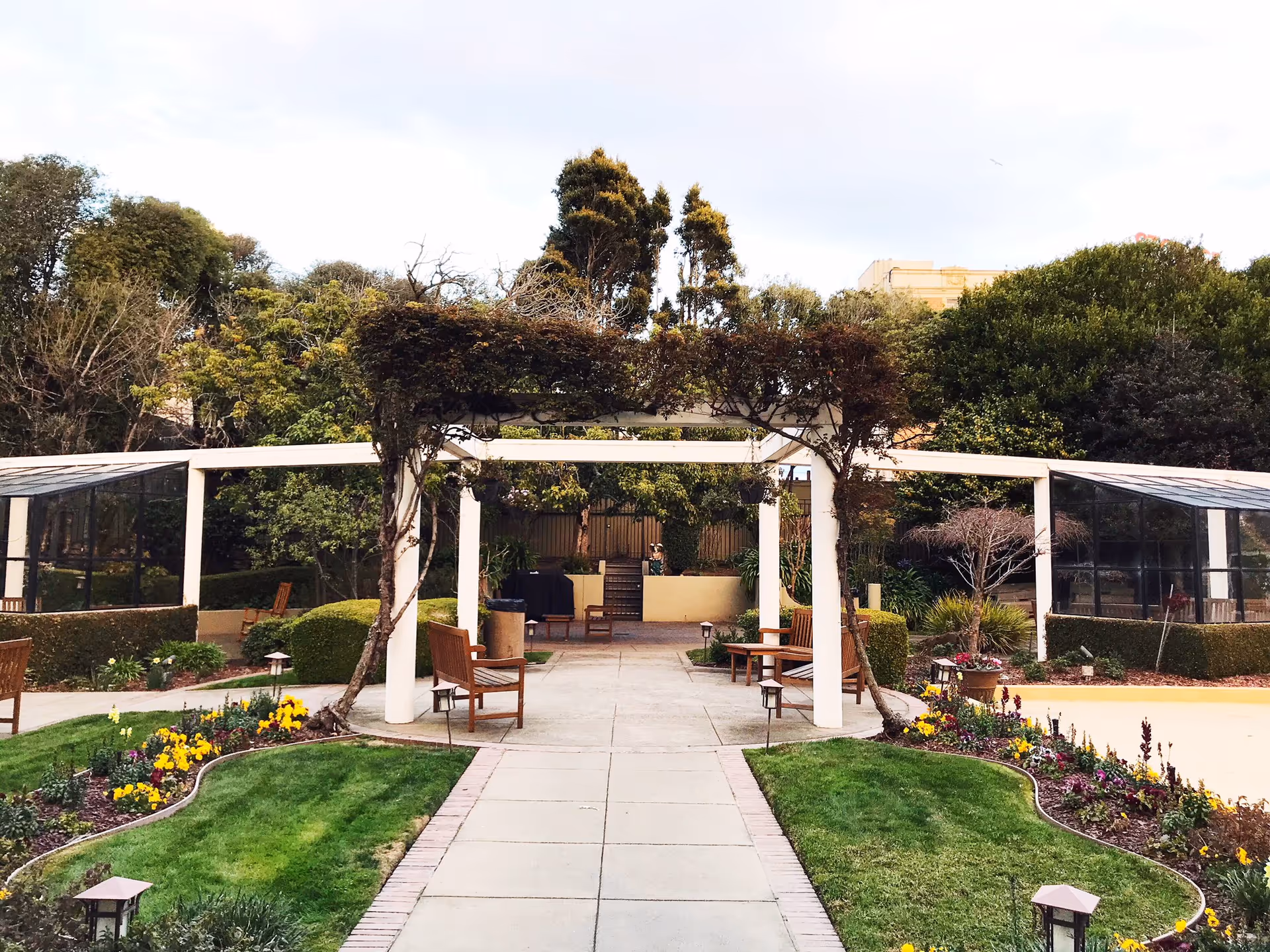 Outdoor garden area with a paved walkway leading to a white pergola covered with climbing plants. There are wooden benches on either side of the walkway and colorful flower beds bordering the grass. Trees and shrubs surround the garden area under a partly cloudy sky.