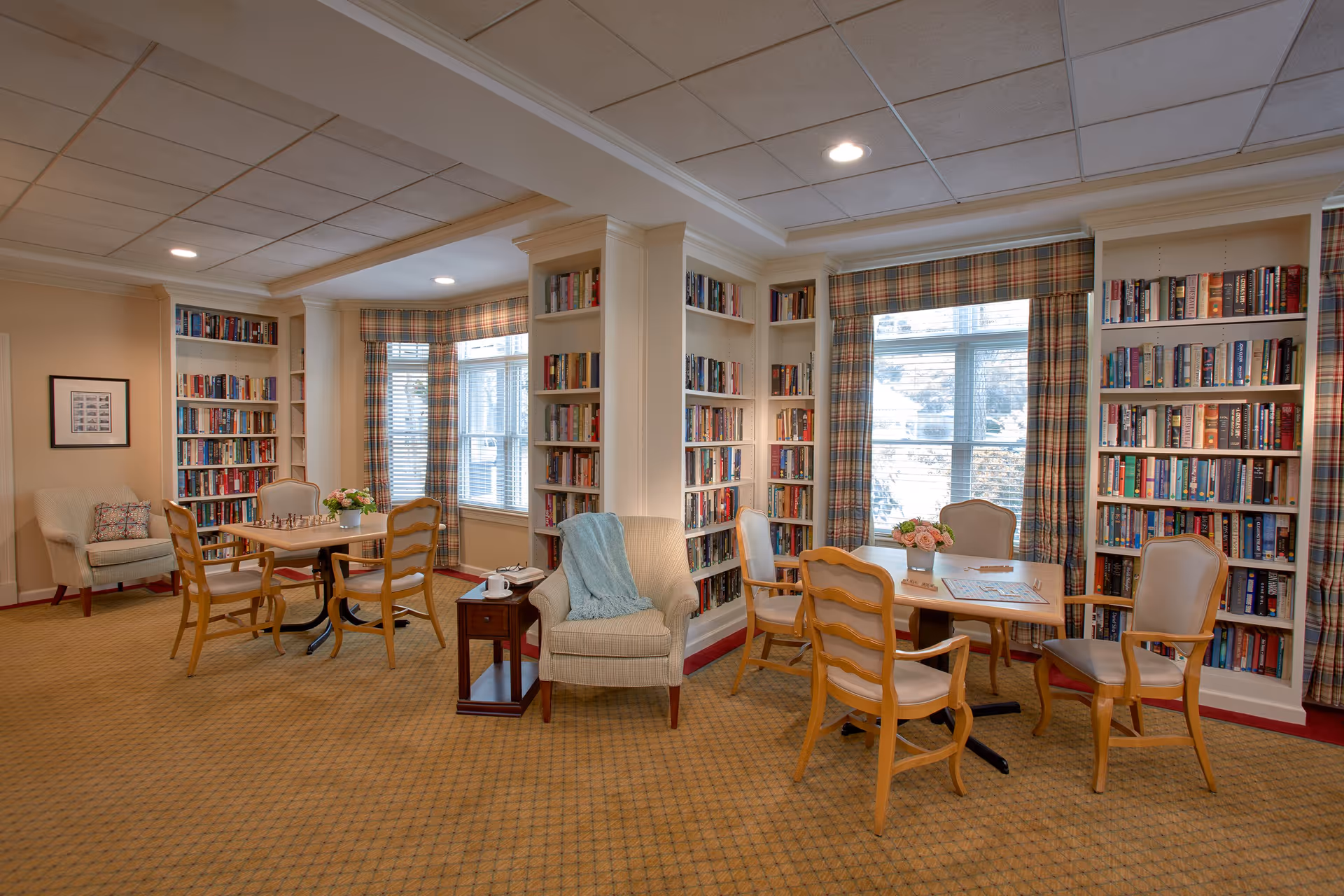 A cozy reading and game area in an assisted living facility with bookshelves filled with books lining the walls. There are two tables with wooden chairs around them, one table has a chess set and the other has a Scrabble game. Comfortable armchairs and side tables with flowers and a cup add to the inviting atmosphere. Large windows with plaid curtains allow natural light to fill the room.