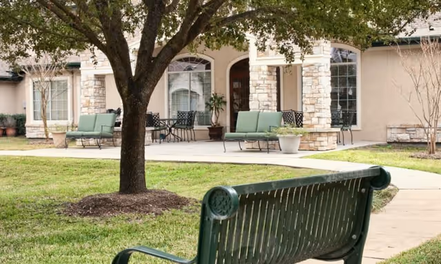 Outdoor seating area with green cushioned chairs and tables on a concrete patio in front of a building with stone pillars and large windows. A tree and a green metal bench are in the foreground on a grassy lawn.