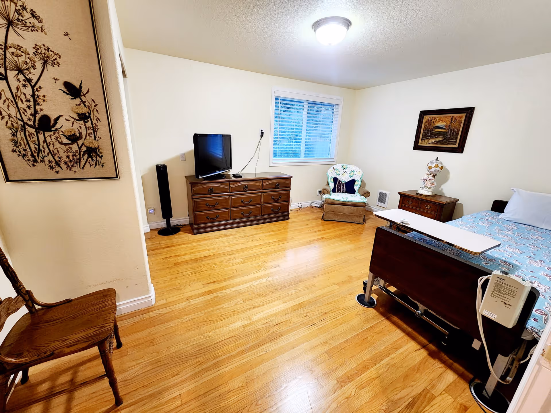 A senior living facility bedroom with a wooden floor, a hospital-style bed with a blue patterned bedspread, a wooden dresser with a TV on top, a cushioned armchair with a floral cover, a wooden side table with a decorative lamp, a wooden chair, and two framed artworks on the walls.