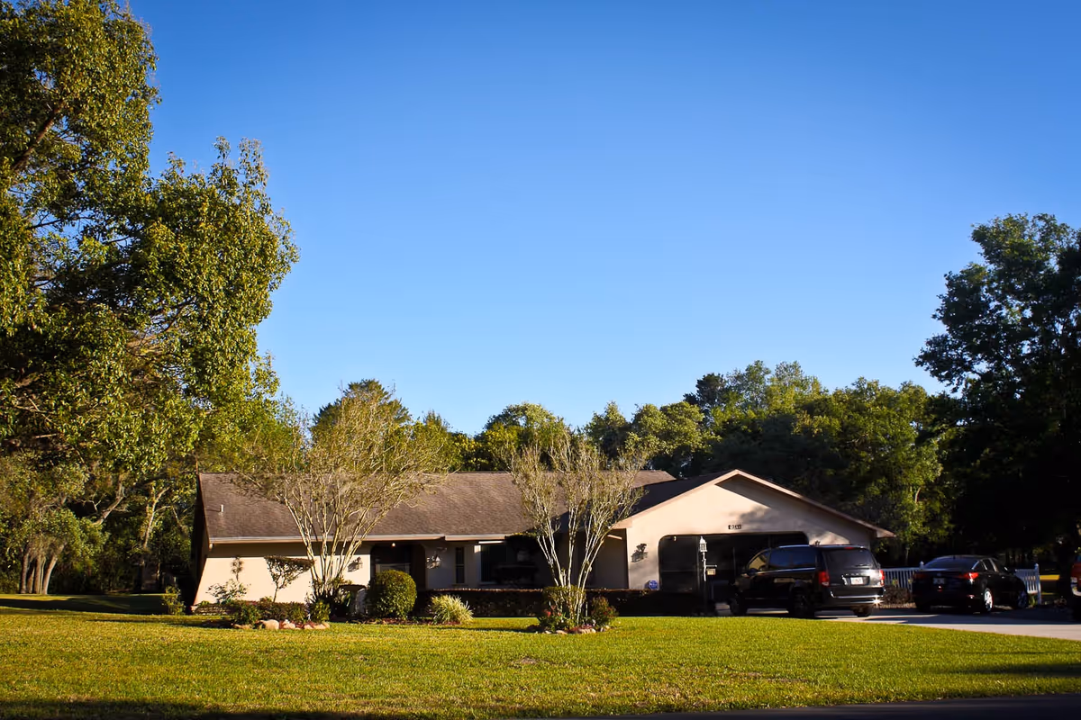 Single-story residential building with a sloped roof surrounded by green trees and a well-maintained lawn under a clear blue sky. Several cars are parked in the driveway to the right of the building.